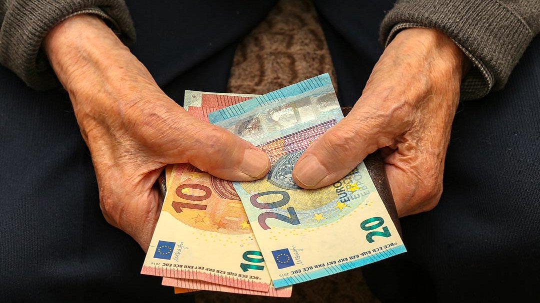 Ältere Hände, die einen Fächer aus Euro-Banknoten halten, darunter 10-, 20- und 50-Euro-Scheine; die Person trägt einen grauen Pullover und eine blaue Hose.