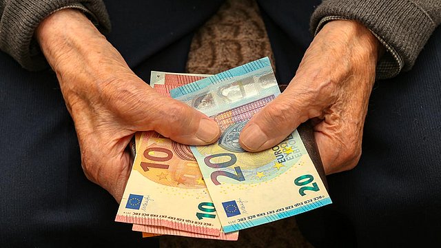 Ältere Hände, die einen Fächer aus Euro-Banknoten halten, darunter 10-, 20- und 50-Euro-Scheine; die Person trägt einen grauen Pullover und eine blaue Hose.