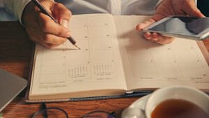 Eine Person, die ein Smartphone in der Hand hält, schreibt mit einem Bleistift an einem Tisch in einen Kalender. Auf dem Tisch stehen außerdem eine Tasse Tee, eine Brille und ein Laptop.
