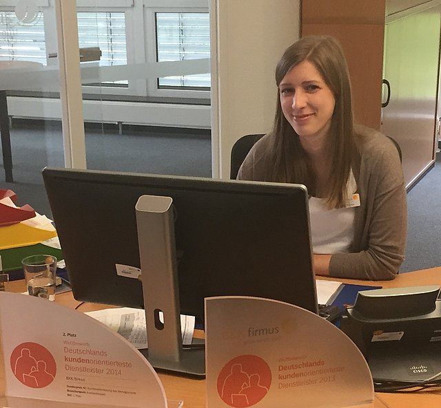 Lisa Marten Eine Frau sitzt an einem Bürotisch mit Auszeichnungen vor sich, arbeitet am Computer und lächelt.