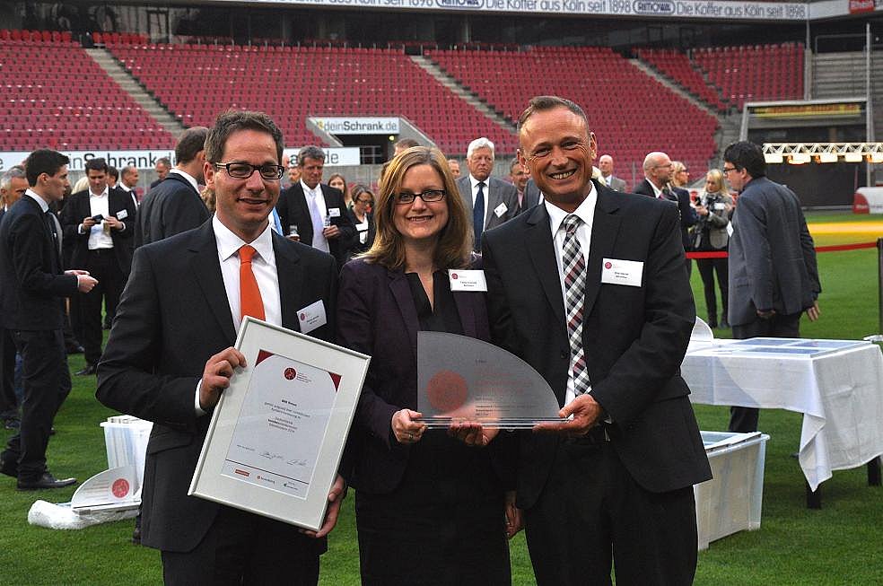 Drei Personen in Geschäftskleidung halten Auszeichnungen und lächeln bei einer Veranstaltung im Freien auf einem Sportplatz.