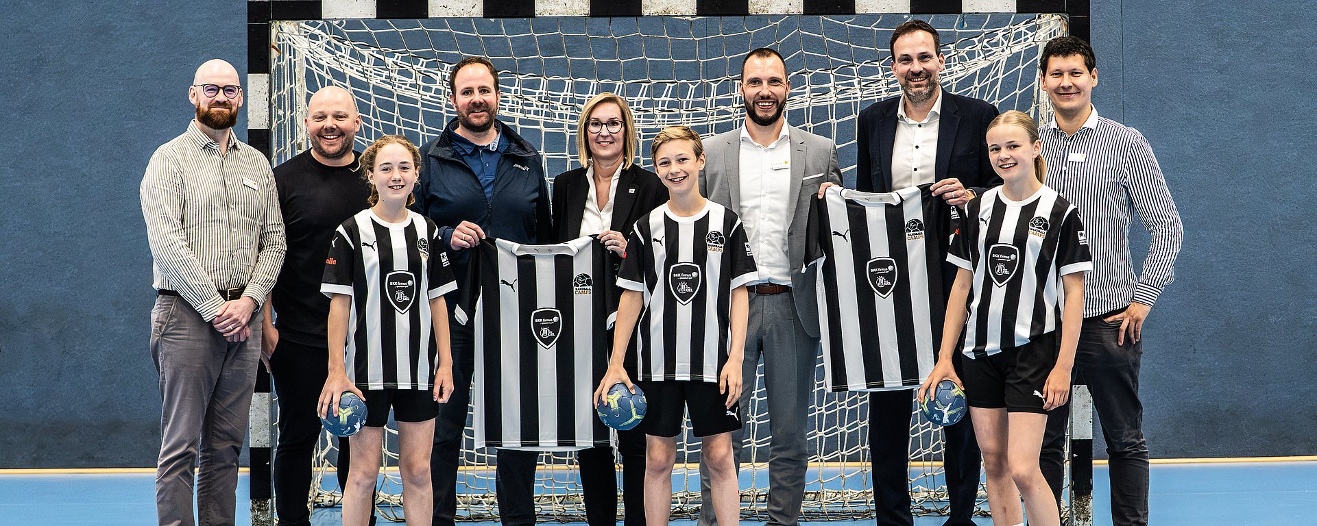 Eine Gruppe aus Erwachsenen und Kindern, die Trikots in den Händen halten, posiert vor einem Handballtor in einer Sporthalle.