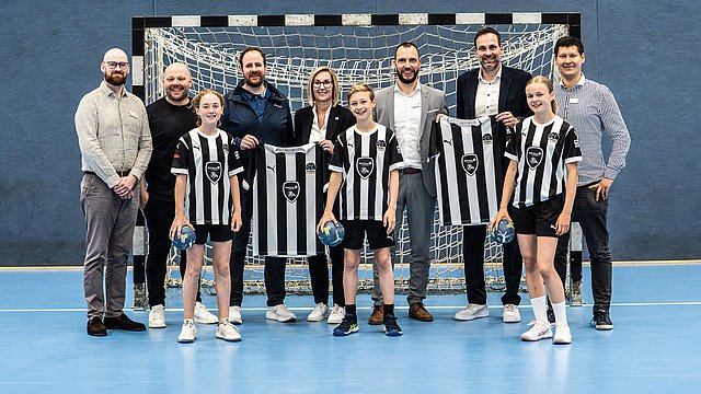 Eine Gruppe aus Erwachsenen und Kindern, die Trikots in den Händen halten, posiert vor einem Handballtor in einer Sporthalle.