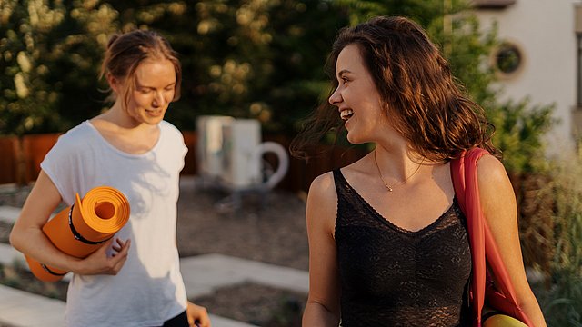 Zwei junge Frauen mit aufgerollter Yoga-Matte in Sportkleidung.
