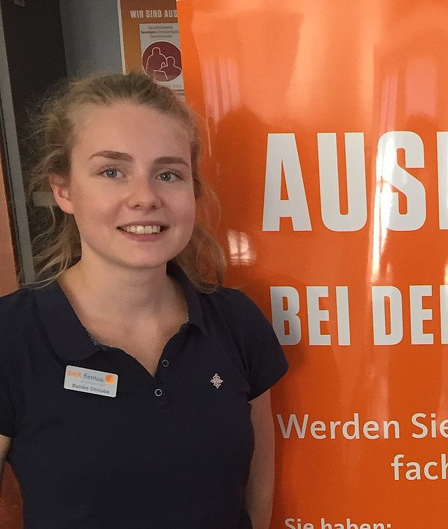 Eunike Dziouba Junge Frau mit blondem Haar steht lächelnd neben einem orangefarbenen Rekrutierungsbanner mit deutschem Text.