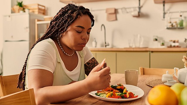 Eine Frau mit geflochtenen Zöpfen sitzt an einem Küchentisch, blickt auf einen Teller mit Essen hinunter und wirkt nachdenklich. Auf dem Tisch und in den Regalen im Hintergrund liegen Obst, eine Tasse und Küchenutensilien.
