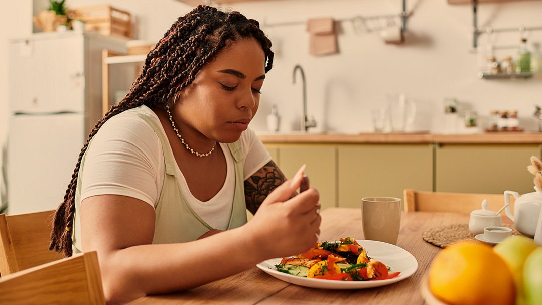 Eine Frau mit geflochtenen Zöpfen sitzt an einem Küchentisch, blickt auf einen Teller mit Essen hinunter und wirkt nachdenklich. Auf dem Tisch und in den Regalen im Hintergrund liegen Obst, eine Tasse und Küchenutensilien.