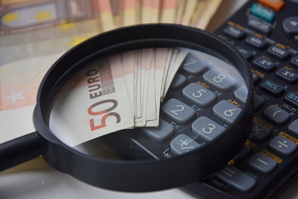 Lupe über 50-Euro-Scheinen und einem Taschenrechner, im Hintergrund weitere Euro-Noten.