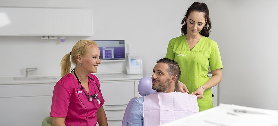 Zwei Zahnärzte unterhalten sich mit einem männlichen Patienten, der in einer hellen Klinik auf einem Zahnarztstuhl sitzt.