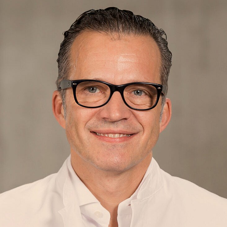 Ein Mann mittleren Alters mit kurzen dunklen Haaren und Brille, der ein weißes Hemd mit Kragen trägt und vor einem schlichten, neutralen Hintergrund in die Kamera lächelt.