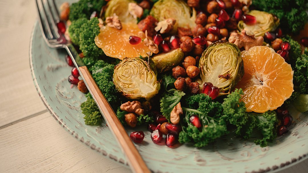 Ein Teller mit Salat aus Grünkohl, geröstetem Rosenkohl, Orangenscheiben, Kichererbsen, Granatapfelkernen und Walnüssen. Eine Gabel liegt am Tellerrand, der auf einem hellen Holztisch steht.