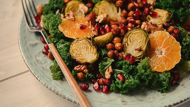 Ein Teller mit Salat aus Grünkohl, geröstetem Rosenkohl, Orangenscheiben, Kichererbsen, Granatapfelkernen und Walnüssen. Eine Gabel liegt am Tellerrand, der auf einem hellen Holztisch steht.
