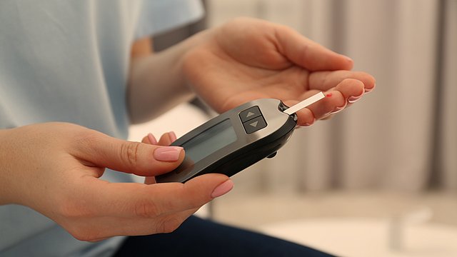 Eine Person hält ein Blutzuckermessgerät mit einem eingelegten Teststreifen in der Hand und misst damit ihren Blutzuckerspiegel. An einer Fingerspitze befindet sich ein kleiner Blutstropfen.