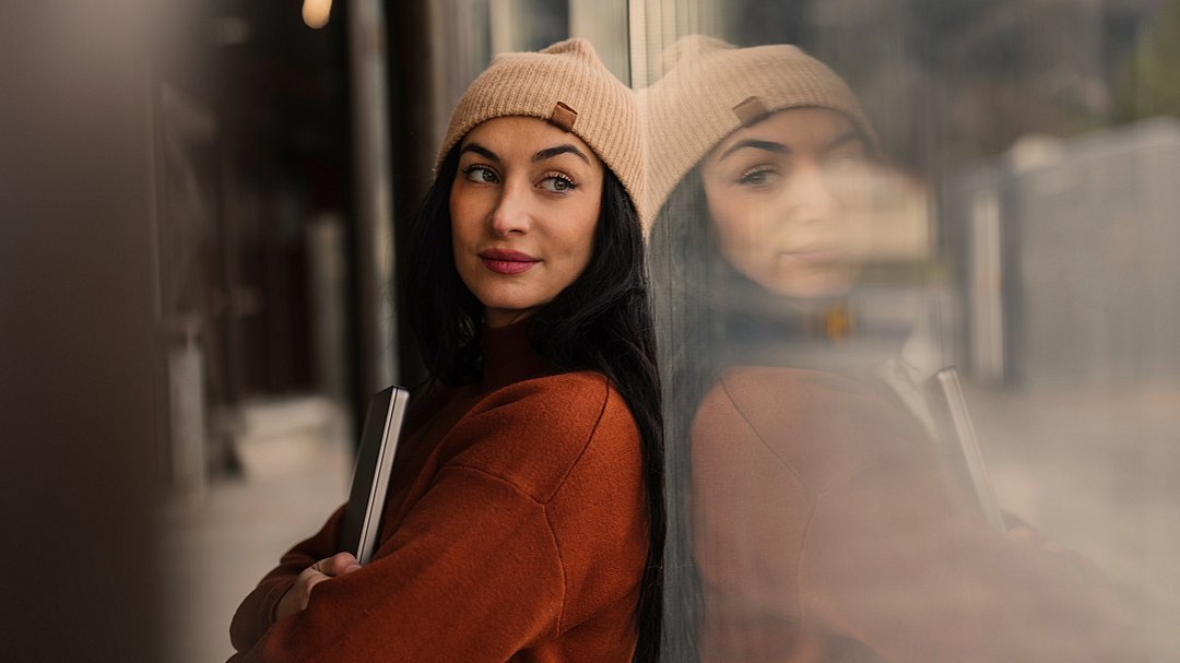 Eine Frau mit einer hellbraunen Mütze und einem braunen Pullover hält einen geschlossenen Laptop in der Hand und lehnt an einer Glaswand, in der sich ihr Spiegelbild widerspiegelt. Sie blickt mit einem leichten Lächeln zur Seite, während sie sich in einer städtischen Außenkulisse befindet.