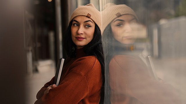 Eine Frau mit einer hellbraunen Mütze und einem braunen Pullover hält einen geschlossenen Laptop in der Hand und lehnt an einer Glaswand, in der sich ihr Spiegelbild widerspiegelt. Sie blickt mit einem leichten Lächeln zur Seite, während sie sich in einer städtischen Außenkulisse befindet.