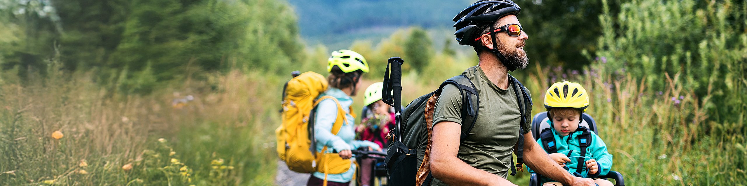 Eine junge Familie in sportlicher Kleidung steht mit Fahrrädern auf einer Wiese, die beiden Kinder sind auf den Fahrrädern im Kindersitz.