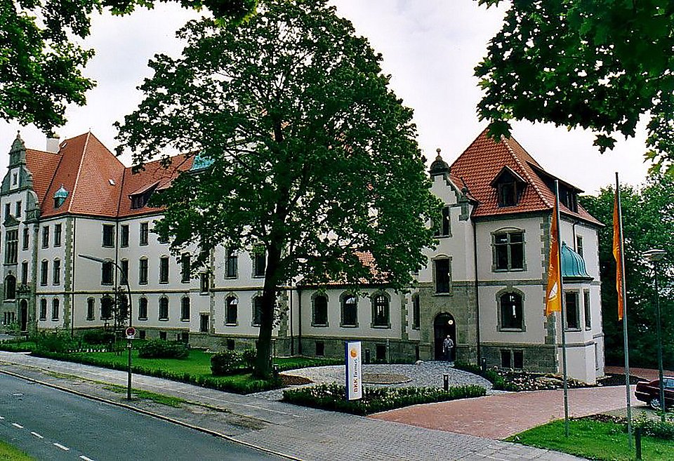Großes, historisches Gebäude mit rotem Ziegeldach, umgeben von Bäumen, Rasen und einer Straße davor.