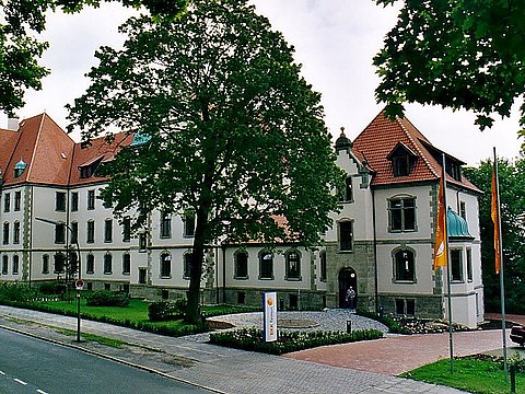 Großes, historisches Gebäude mit rotem Ziegeldach, umgeben von Bäumen, Rasen und einer Straße davor.