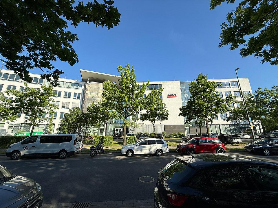 Standort Essen Modernes Bürogebäude mit meinestadt.de-Schild, parkende Autos entlang der Straße, Bäume rahmen die Aussicht ein.