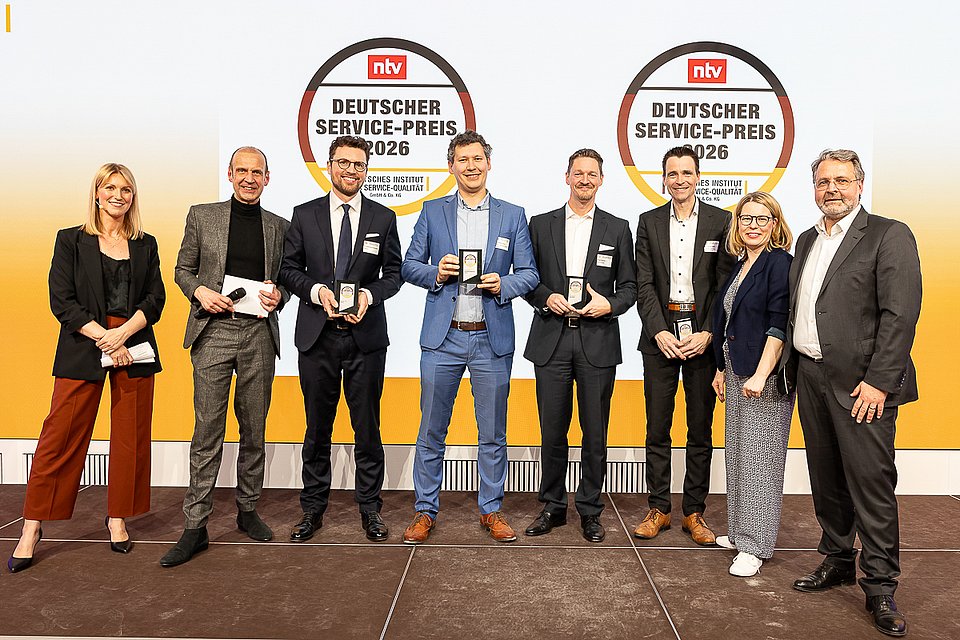Acht Personen stehen auf der Bühne des Deutschen Service-Preises 2026 und halten ihre Preise in den Händen. Sie lächeln für ein Foto.