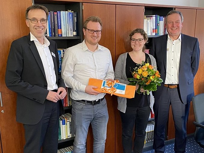 Vier lächelnde Erwachsene stehen in einem Haus; einer hält Blumen, ein anderer eine Geschenkkarte. Der Text in deutscher Sprache ist überlagert.