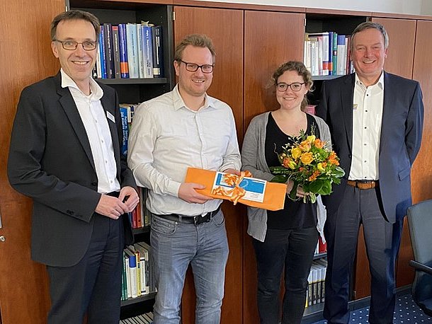 Vier lächelnde Erwachsene stehen in einem Haus; einer hält Blumen, ein anderer eine Geschenkkarte. Der Text in deutscher Sprache ist überlagert.