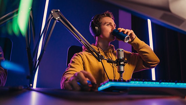 Eine Person mit Kopfhörern sitzt an einem Schreibtisch mit Mikrofon und Tastatur und trinkt aus einer Dose in einem neonbeleuchteten Raum; wahrscheinlich spielt sie gerade ein Videospiel oder streamt online.