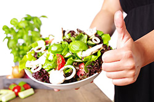 Person, die eine Schüssel mit frischem Salat hält und den Daumen nach oben streckt, mit Gemüse im Hintergrund.