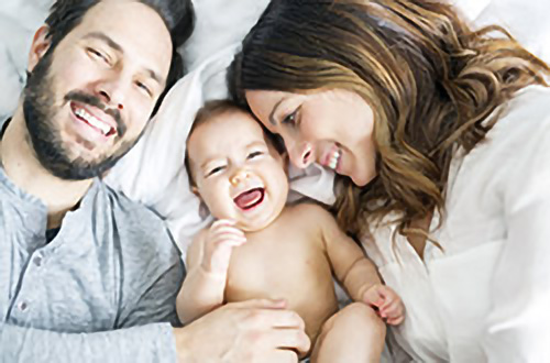 Ein lächelnder Mann und eine lachende Frau liegen im Bett, zwischen ihnen ein lachendes Baby.