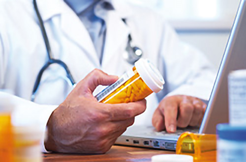 Ein Arzt hält ein Fläschchen mit verschreibungspflichtigen Tabletten in der Hand, während er an einem Schreibtisch einen Laptop benutzt.