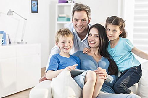 Eine lächelnde vierköpfige Familie, zwei Eltern und zwei Kinder, die in einem hellen Raum auf einer weißen Couch zusammensitzen.