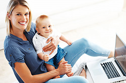 Eine lächelnde Frau mit einem Baby sitzt auf dem Boden neben einem geöffneten Laptop.