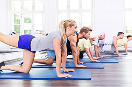 Teilnehmer eines Fitnesskurses machen Bodenübungen auf Matten in einem hellen Raum mit großen Fenstern.