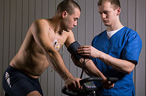 Ein Mann auf einem Heimtrainer wird von einer medizinischen Fachkraft in einem blauen Kittel auf seinen Blutdruck überprüft, wobei Drähte an der Brust befestigt sind.