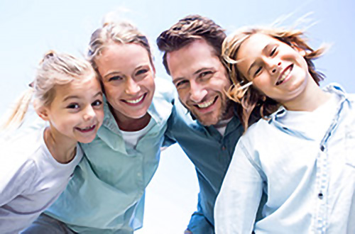 Eine lächelnde vierköpfige Familie, zwei Erwachsene und zwei Kinder, posiert eng beieinander im Freien unter blauem Himmel.