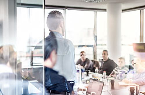 Ein Mann hält eine Präsentation vor einer Gruppe von Personen, die in einem hellen Büro um einen Konferenztisch sitzen.