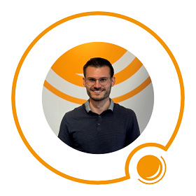 Ein lächelnder Mann mit Brille steht vor einem orange-weißen kreisförmigen Logohintergrund.