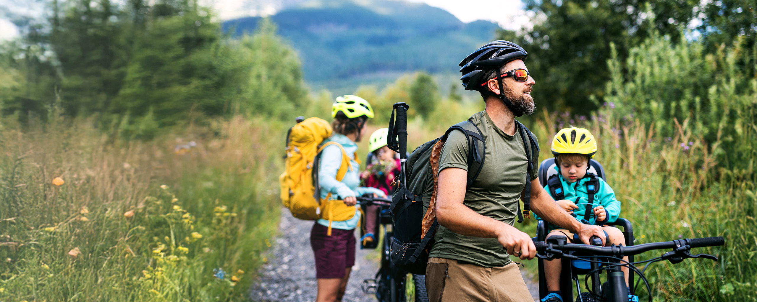 Eine junge Familie in sportlicher Kleidung steht mit Fahrrädern auf einer Wiese, die beiden Kinder sind auf den Fahrrädern im Kindersitz.