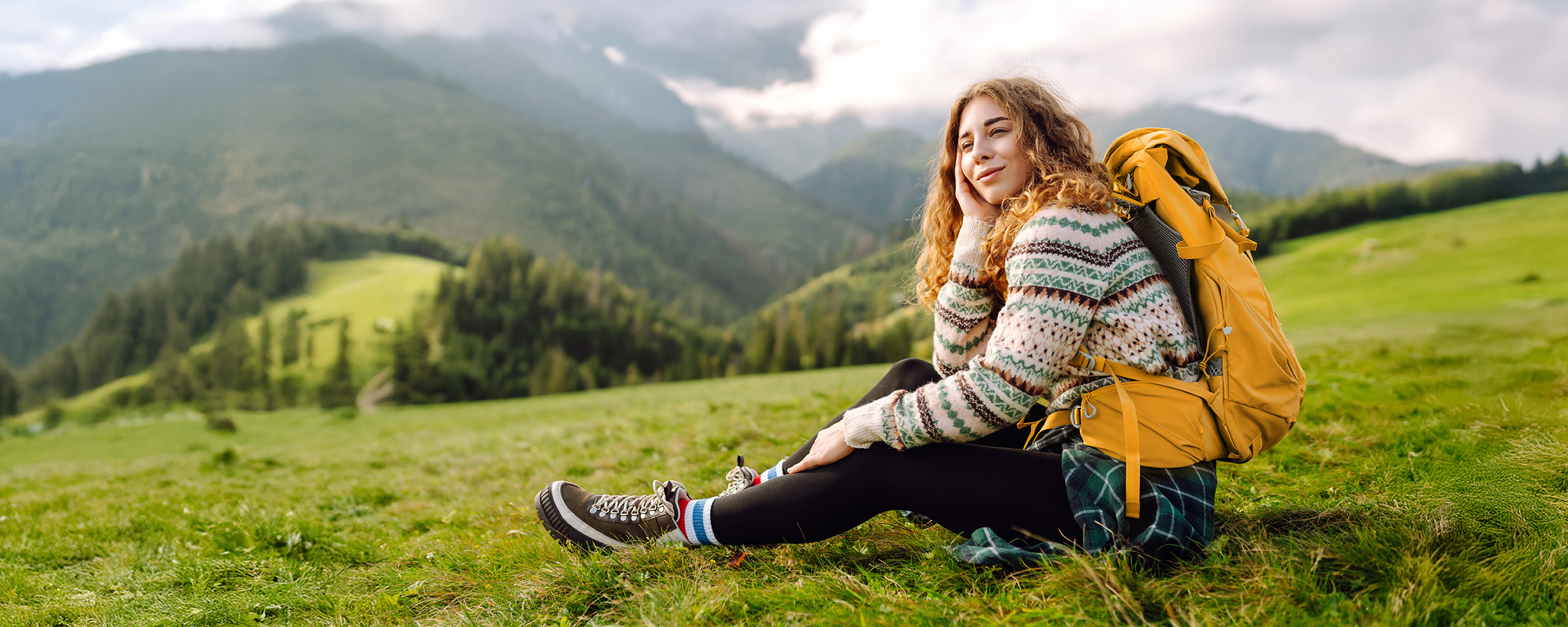 Eine junge Frau mit Rucksack und Wanderstiefeln sitzt auf einer Wiese in den Bergen, sie lächelt. 