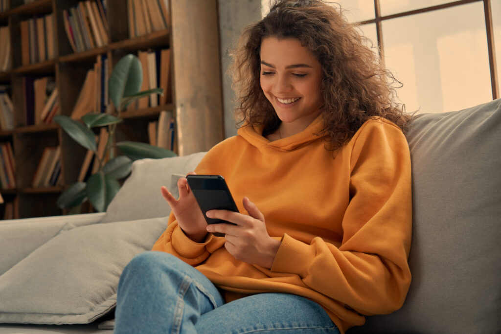 Eine Frau mit lockigem Haar, die einen gelben Kapuzenpulli und Jeans trägt, sitzt lächelnd auf einem Sofa und nutzt ihr Smartphone. Im Hintergrund ist ein Bücherregal zu sehen.