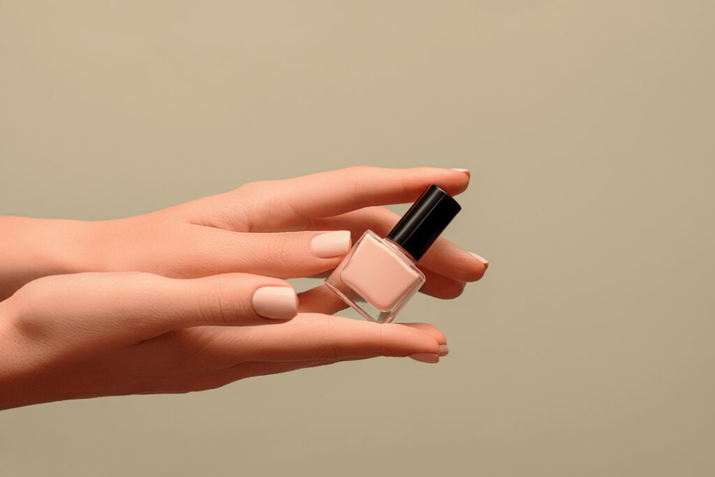 Eine Person mit gepflegten Nägeln hält vor einem schlichten beigen Hintergrund ein kleines Fläschchen mit hellrosa Nagellack in der Hand.