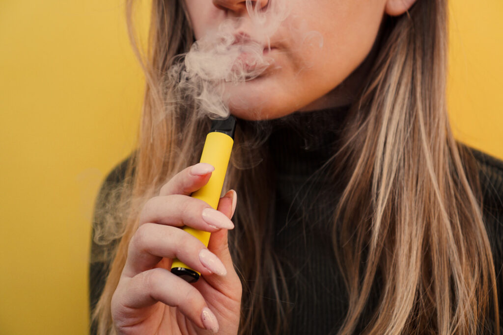 Eine Person mit langen blonden Haaren hält einen gelben Vape-Pen an den Mund und atmet Dampf aus. Der Hintergrund ist einheitlich gelb, und nur die untere Hälfte ihres Gesichts ist zu sehen.