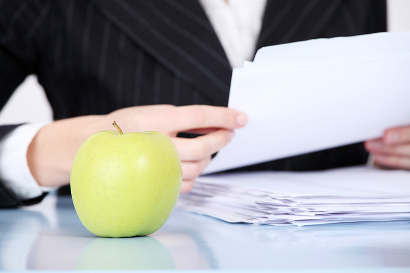 Ein grüner Apfel auf einem Schreibtisch vor einer Person, die einen Stapel Papiere hält und einen dunklen Nadelstreifenanzug trägt.